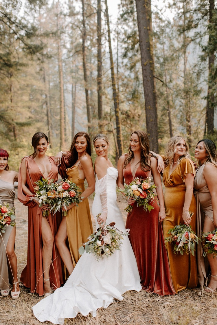 A bride with her bridesmaids in rust and gold dresses among the pines