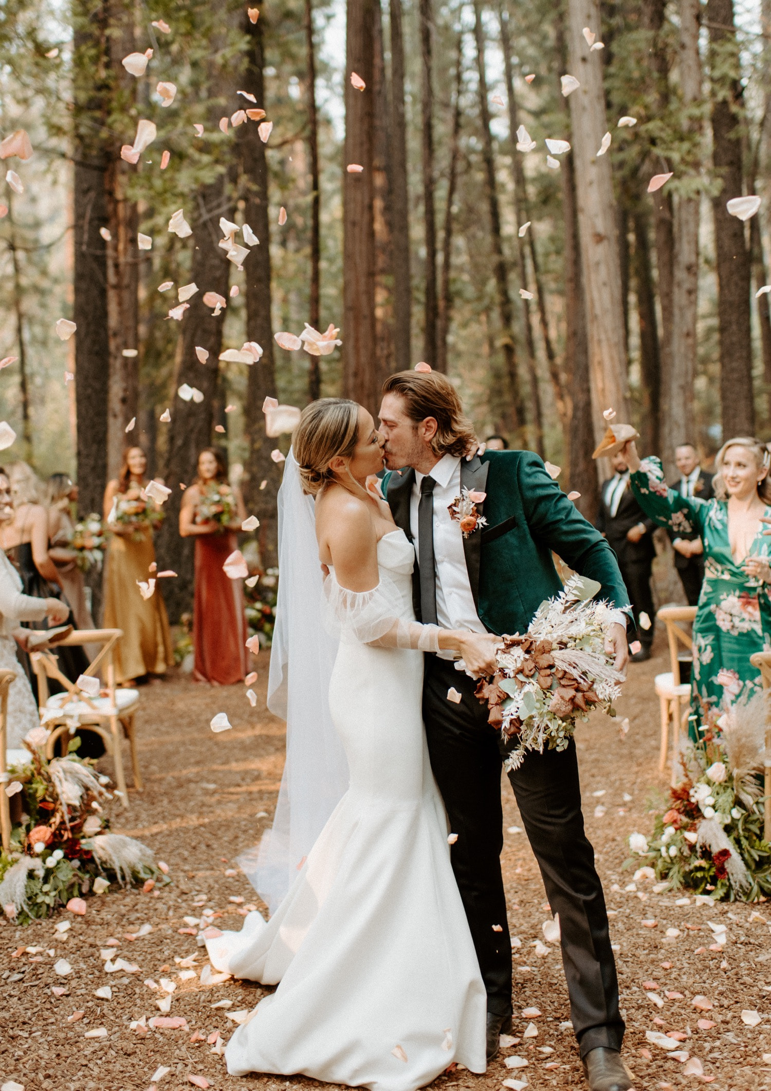 A couple walks back up a forest aisle through a shower of flower petals