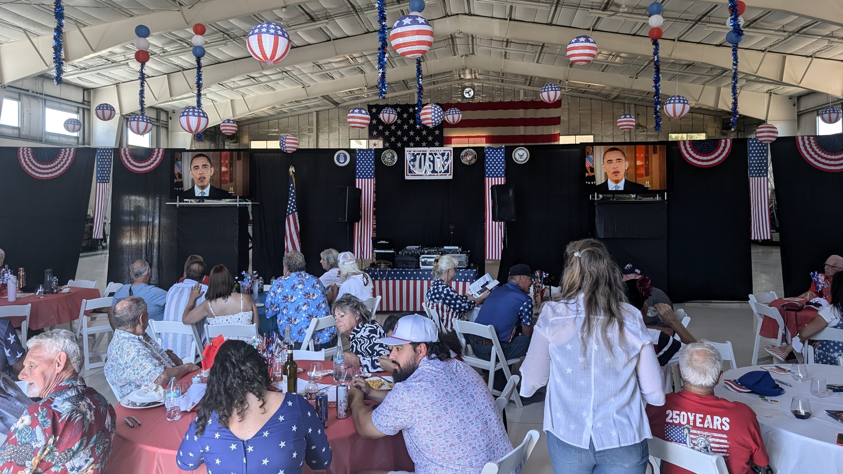 Guests at red, white and blue tables facing the USO stage in an aircraft hangar decorated with flags and star spangled lanterns