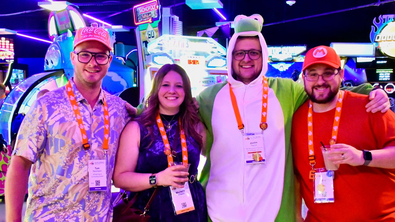 Four AMUG guests posing together at the arcade, one in a green dinosaur onesie