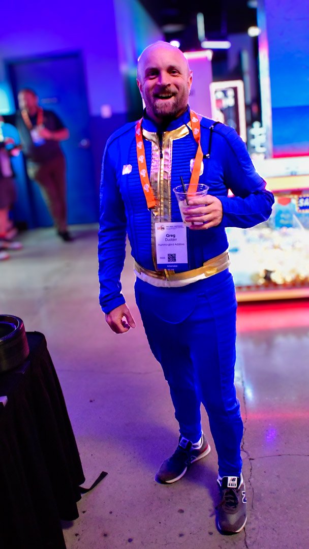 A smiling AMUG guest in a blue and gold jumpsuit with a drink
