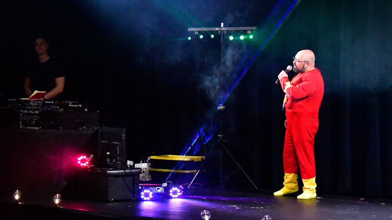An AMUG guest in a red superhero suit singing karaoke on the White Rabbit stage beside the DJ booth, under lasers and fog