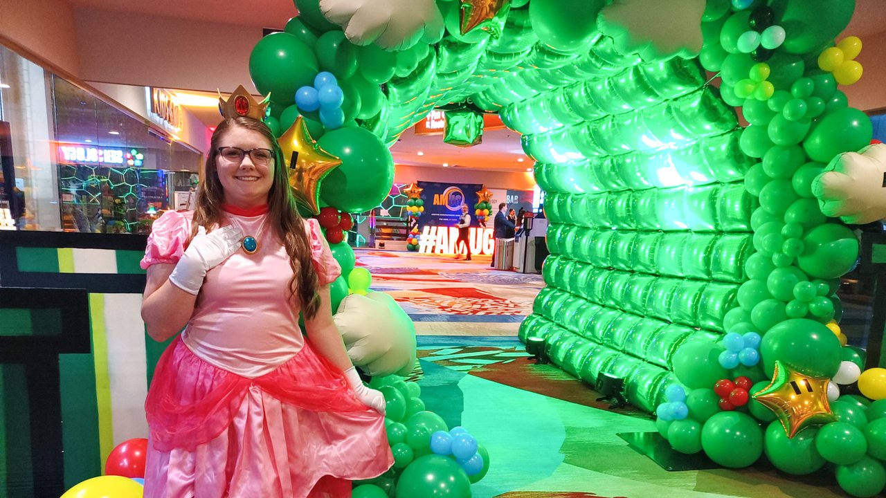 An AMUG guest dressed as Princess Peach smiling in front of the green balloon arch