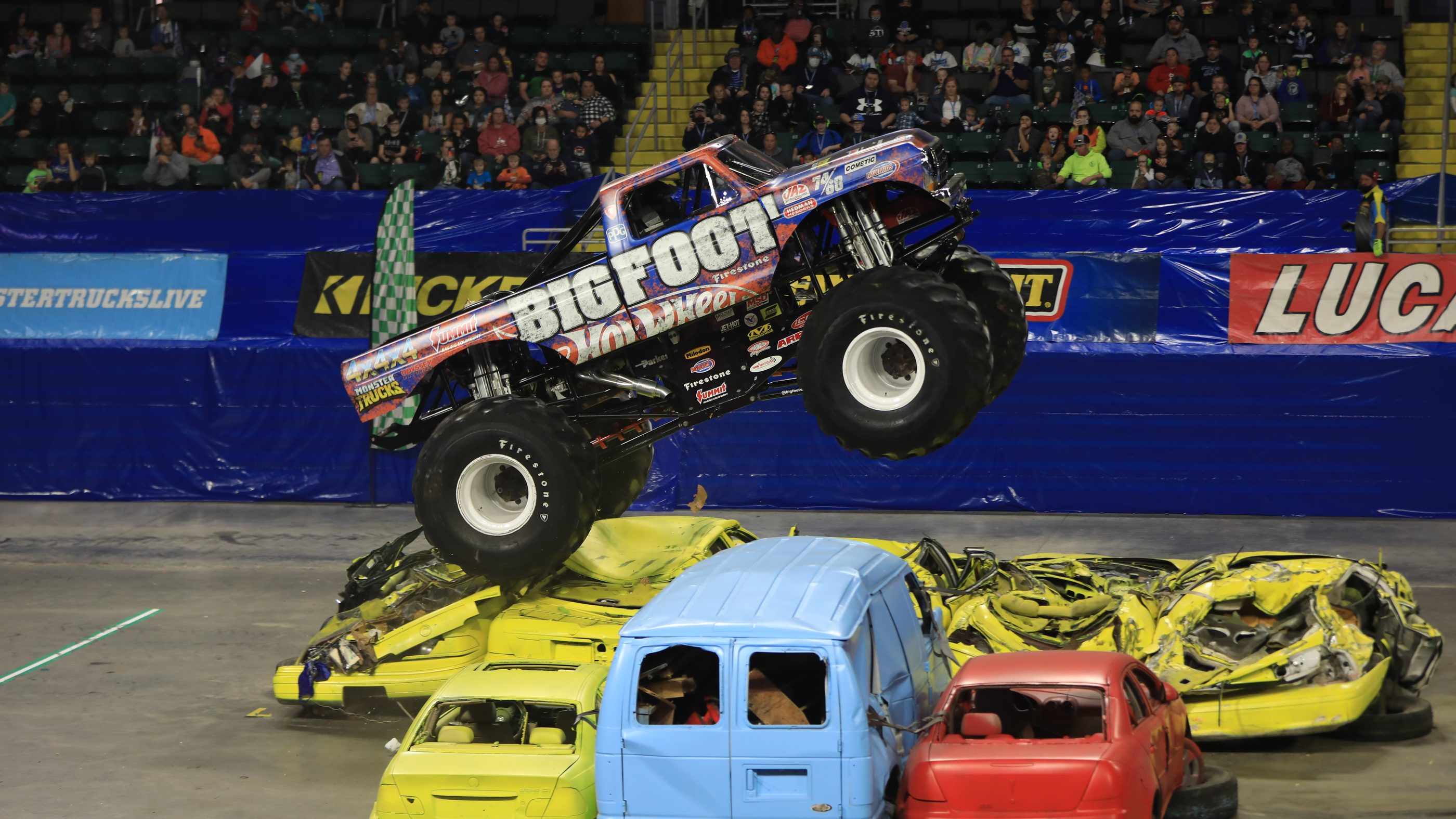 Bigfoot in the air over a row of crushed cars in front of the stands at a KICKER Monster Truck Show