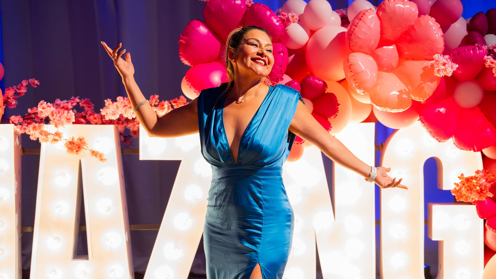 A Renown guest in a blue satin dress throwing her arms wide at the lit AMAZING letters and the balloon arch