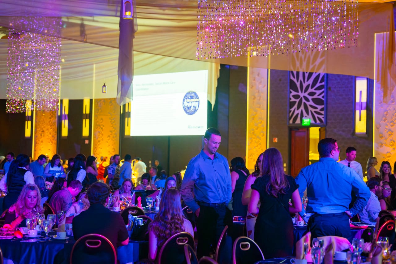 Renown guests mingling between tables as the program plays on the screens