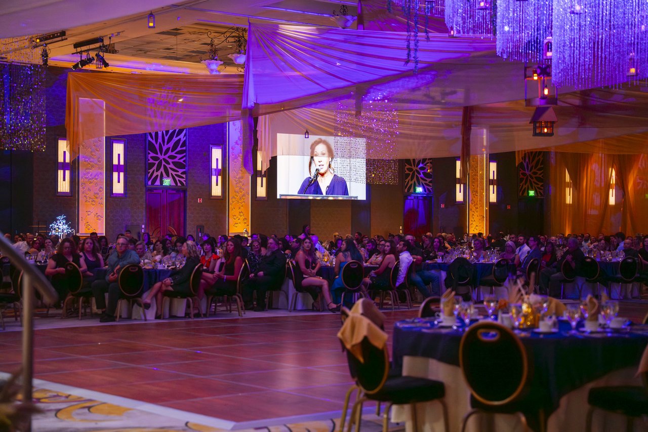 The packed ballroom under purple lights with a speaker on the screen