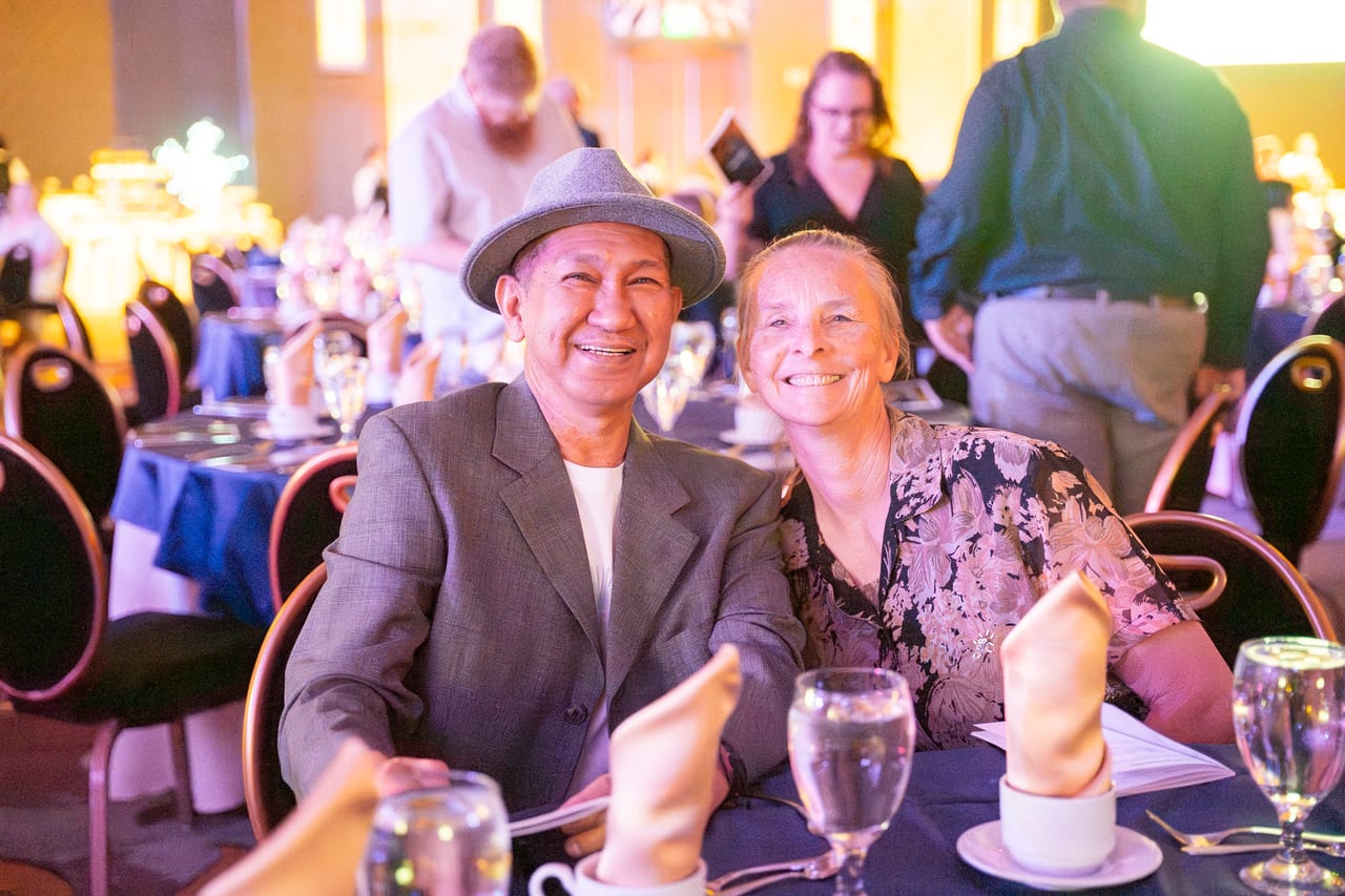 A Renown couple smiling at dinner, him in a gray fedora