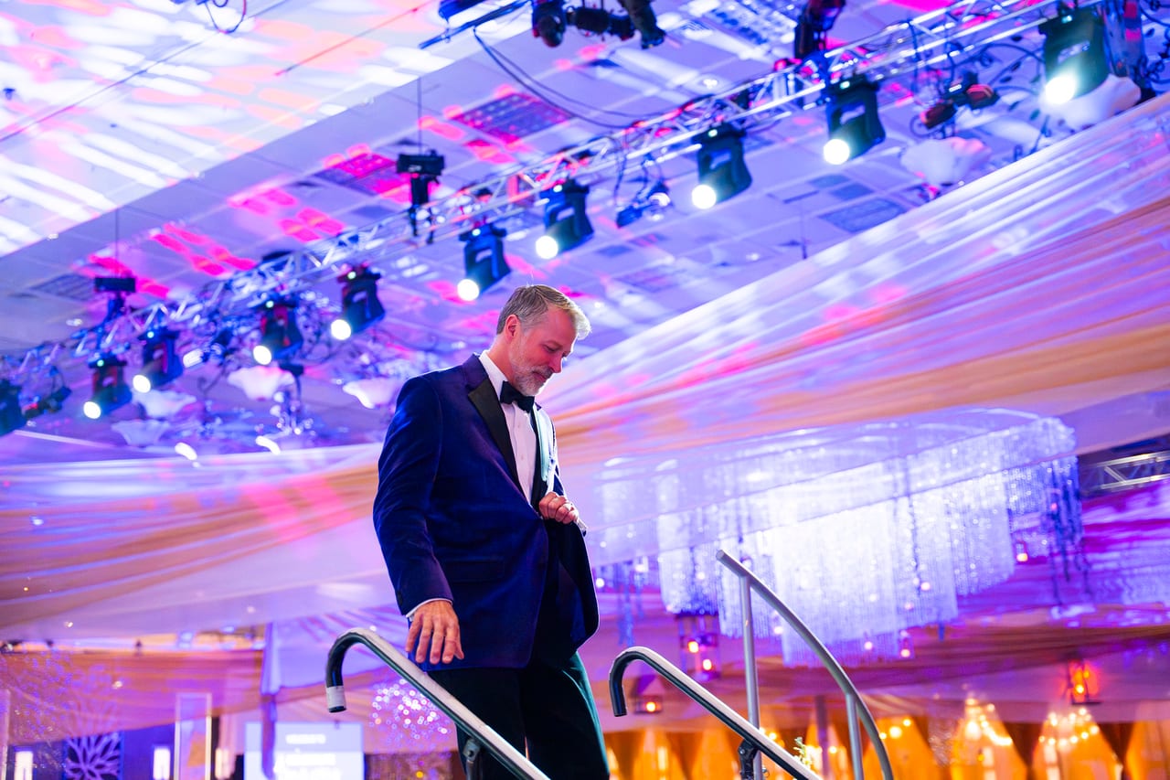 The emcee in a tuxedo heading up the stage steps under the moving lights