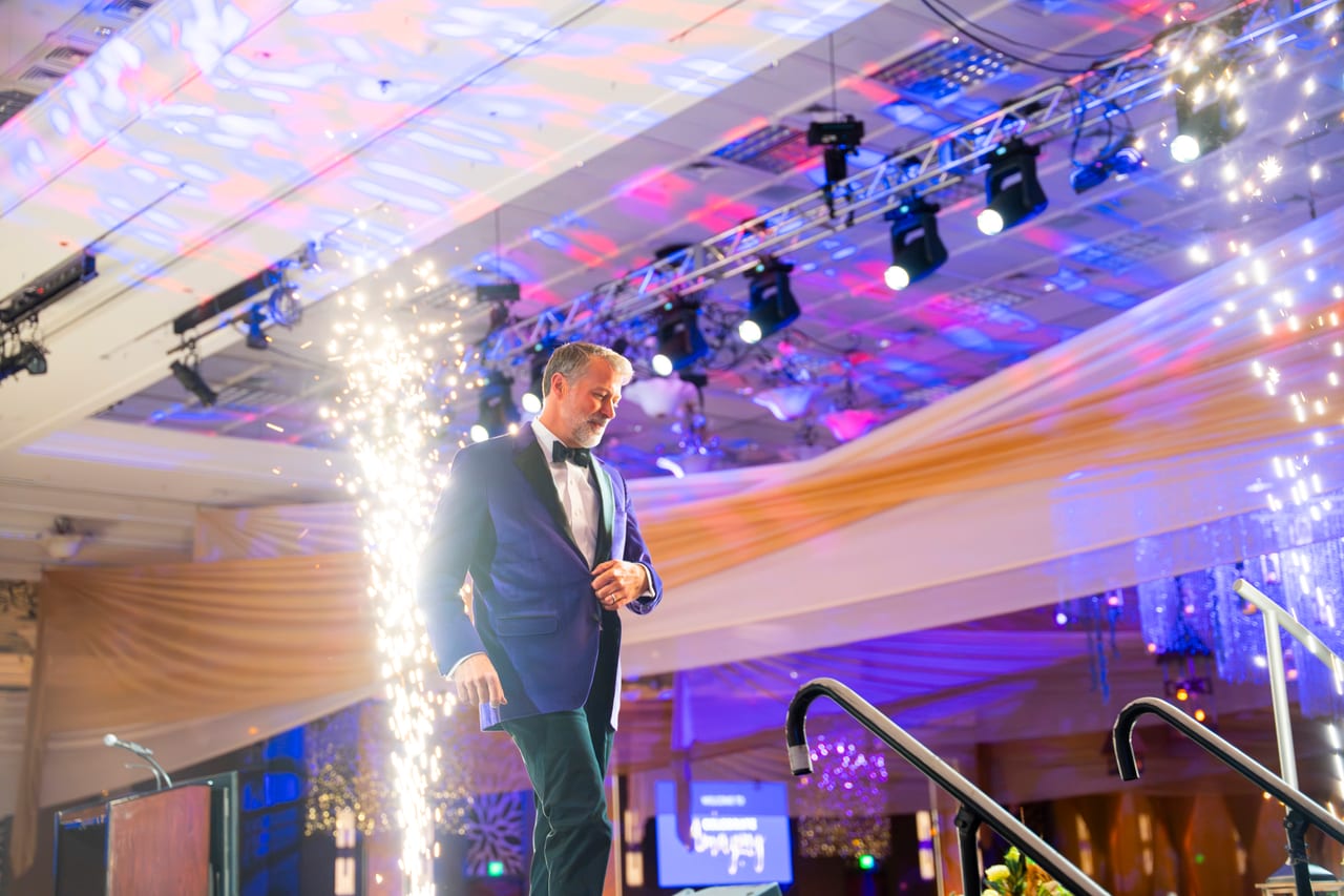 The emcee walking across the stage past a fountain of cold sparks