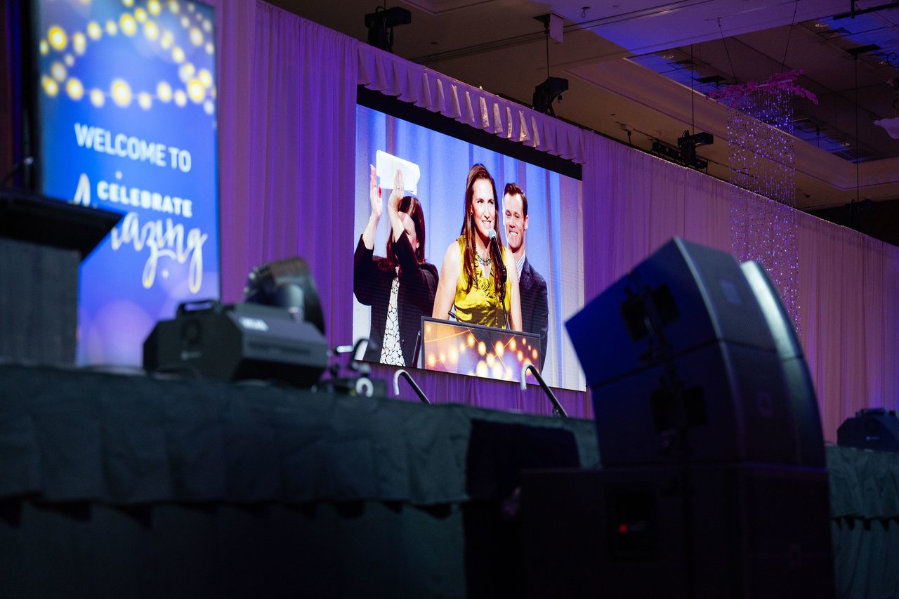 The big screen showing an honoree lifting her award beside the welcome sign