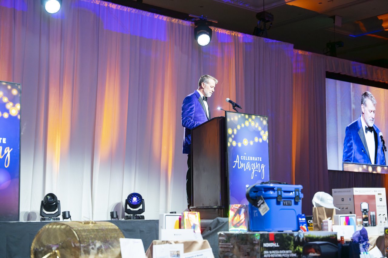 The host speaking beside the gold raffle drum and prizes