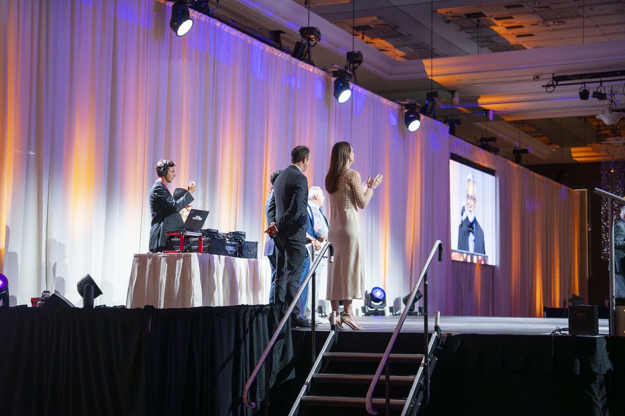 Renown leaders on stage applauding as a speaker appears on the big screen