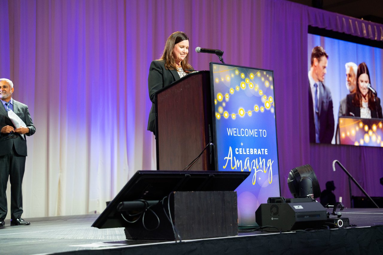 A presenter at the podium with the big screen showing her beside her