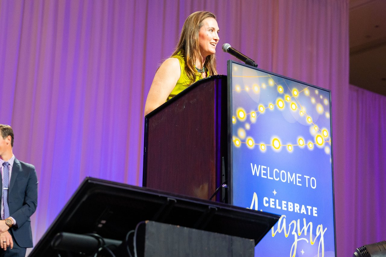 A presenter in a yellow dress speaking at the Celebrate Amazing podium