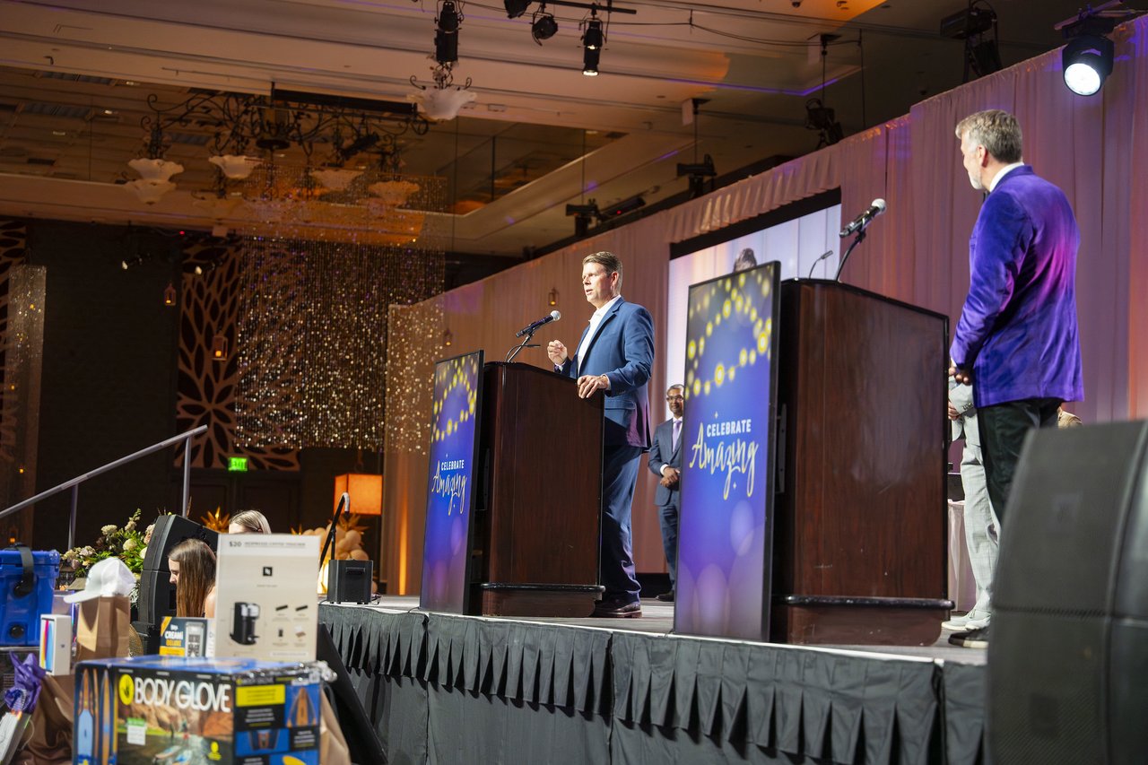 The host watching as a Renown leader speaks at the podium