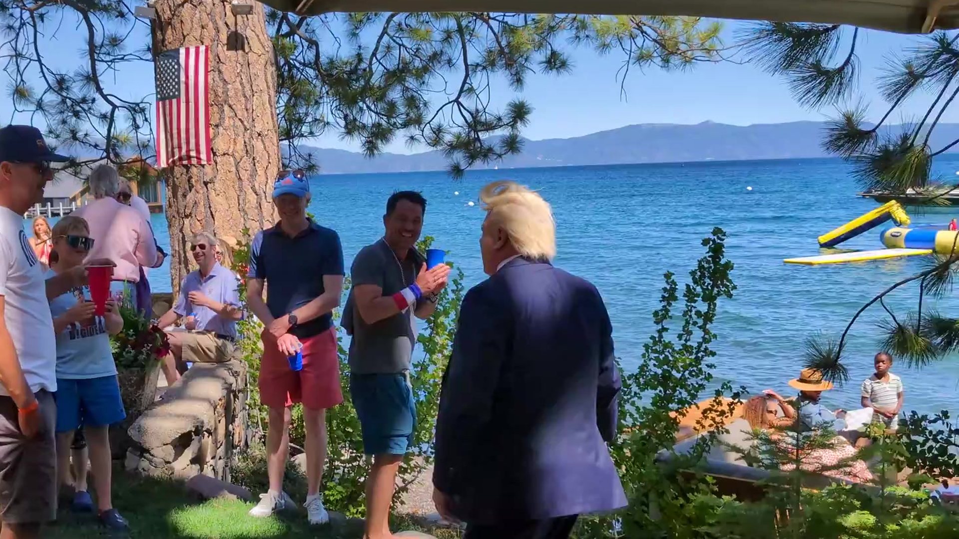 A Trump impersonator in a navy suit greets smiling guests under the American flag on the pine by Lake Tahoe at a Fourth of July party in Tahoe City