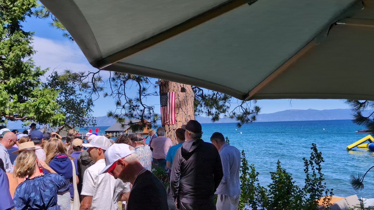 Guests in caps and white shirts by the lake under the flag pine
