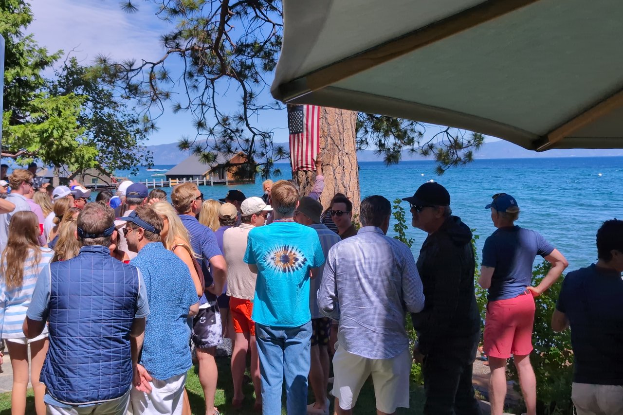 A crowd gathers on the lawn under the flag on the pine, facing the lake