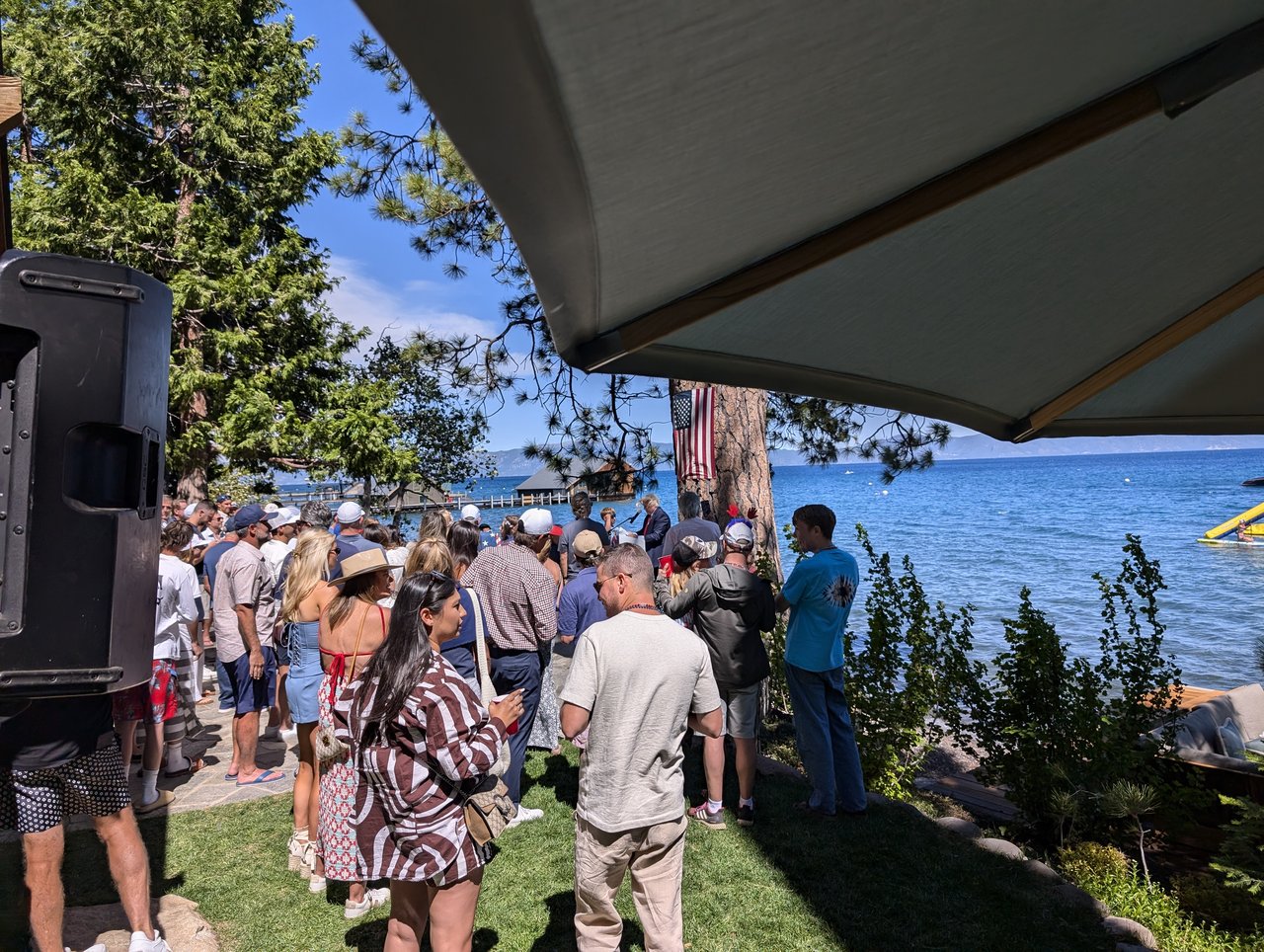 The crowd on the grass by the lake seen past the DJ speaker, a guest in a striped dress