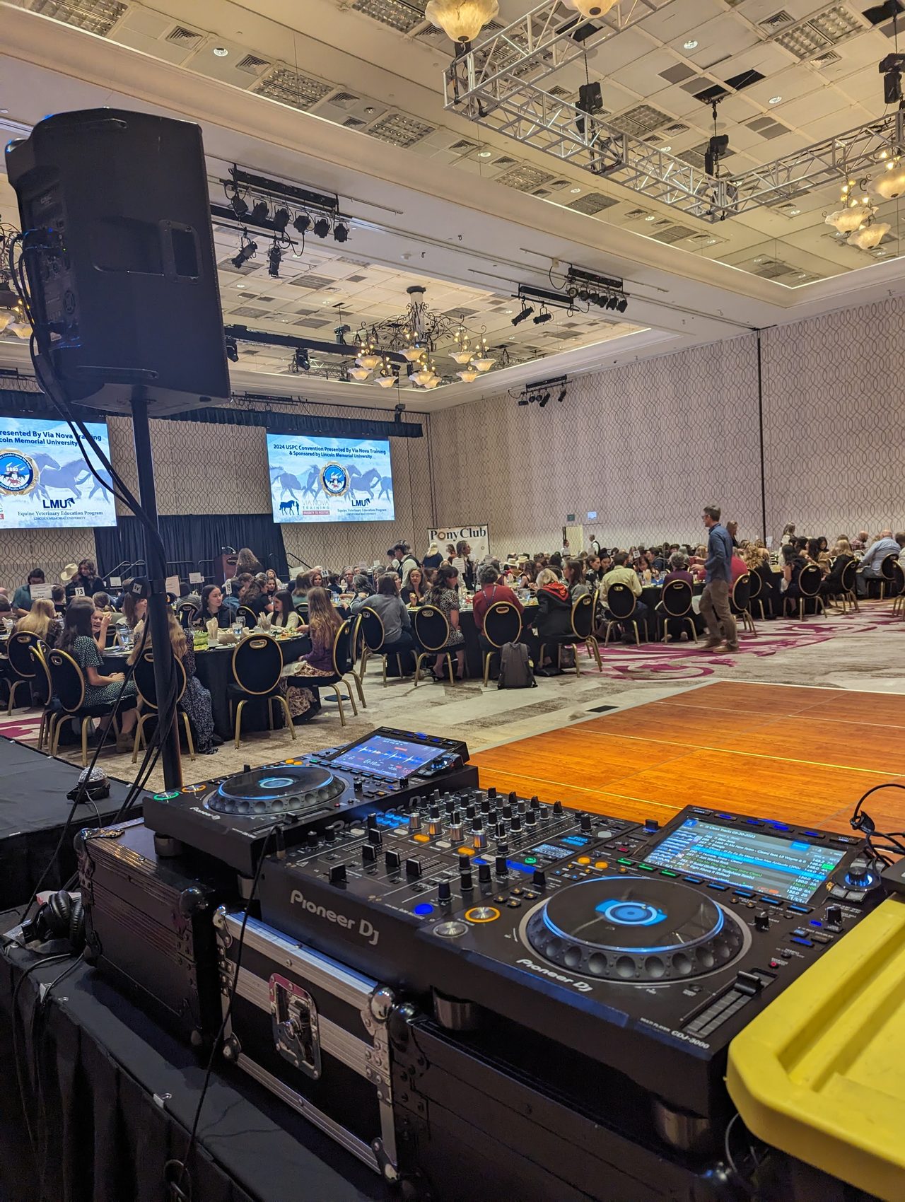 Guests at dinner seen from the DJ booth, with the Pony Club banner and the convention screens behind them