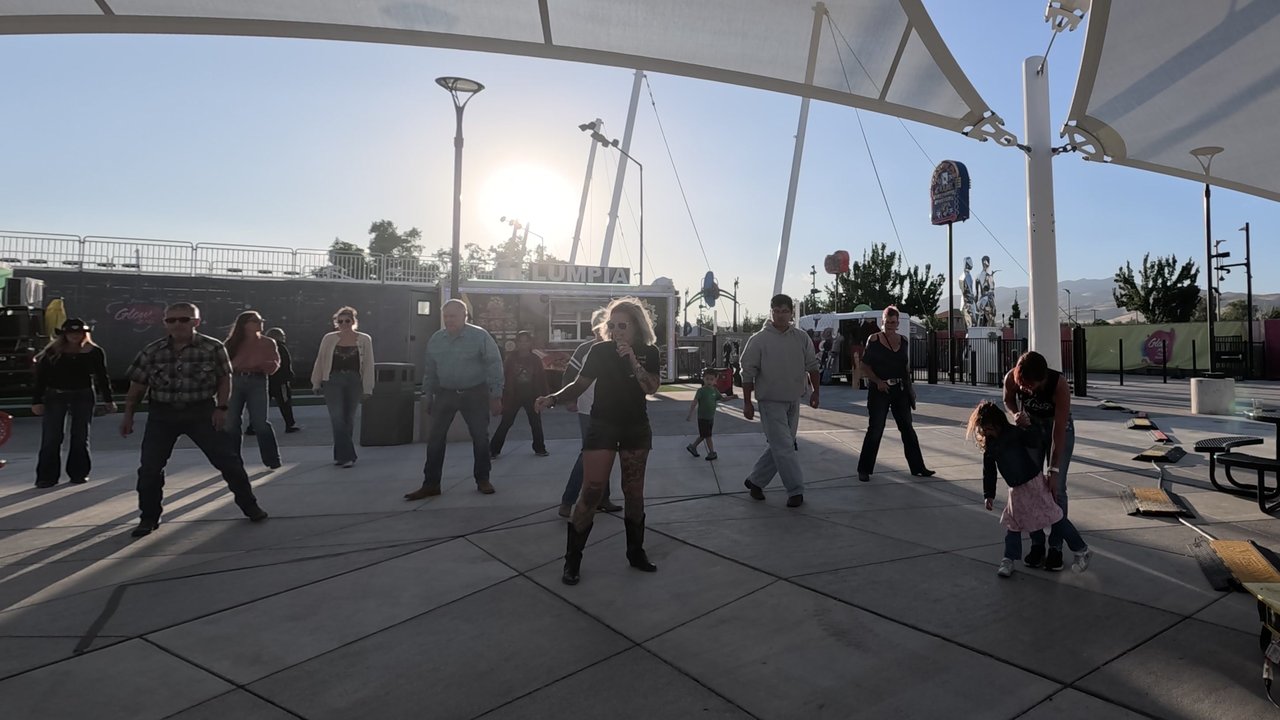 A line dance instructor with a headset mic leads a full floor of dancers in the evening sun