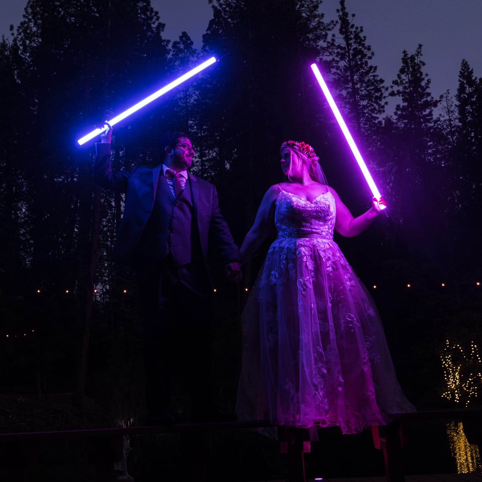 Newlyweds Abney and Chris hold glowing blue and purple lightsabers high in front of tall pines at dusk