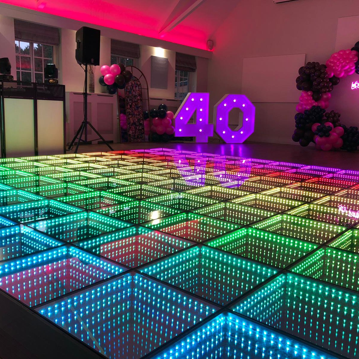 A rainbow lit LED infinity dance floor fills a party hall, with the DJ booth on the left and a balloon arch at the back