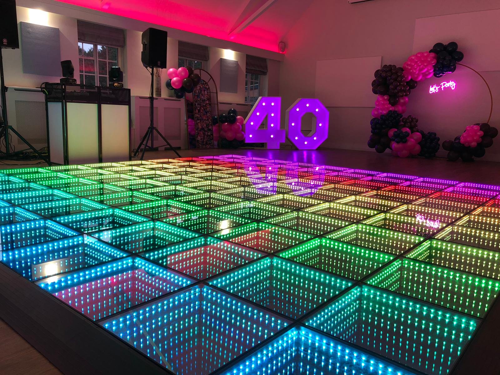 A rainbow lit LED infinity dance floor fills a party hall set for a 40th birthday, with the DJ booth on the left and a balloon arch at the back