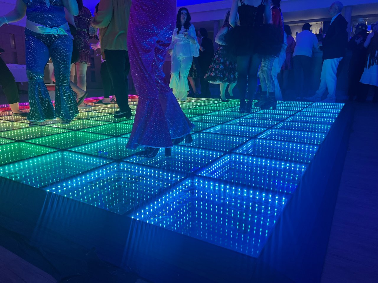 Guests in disco costumes dance on a raised LED infinity dance floor glowing green and blue