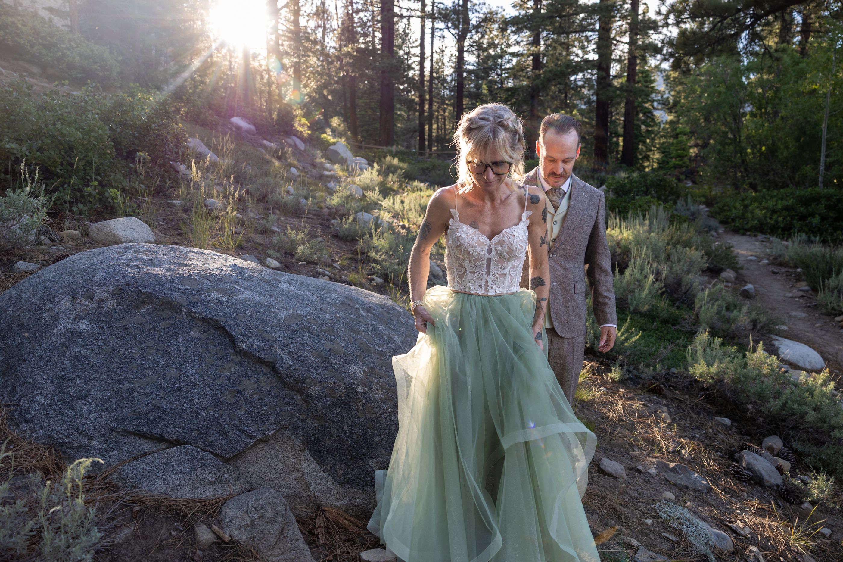 Amy lifting her sage green wedding dress on a forest trail as Justin follows behind