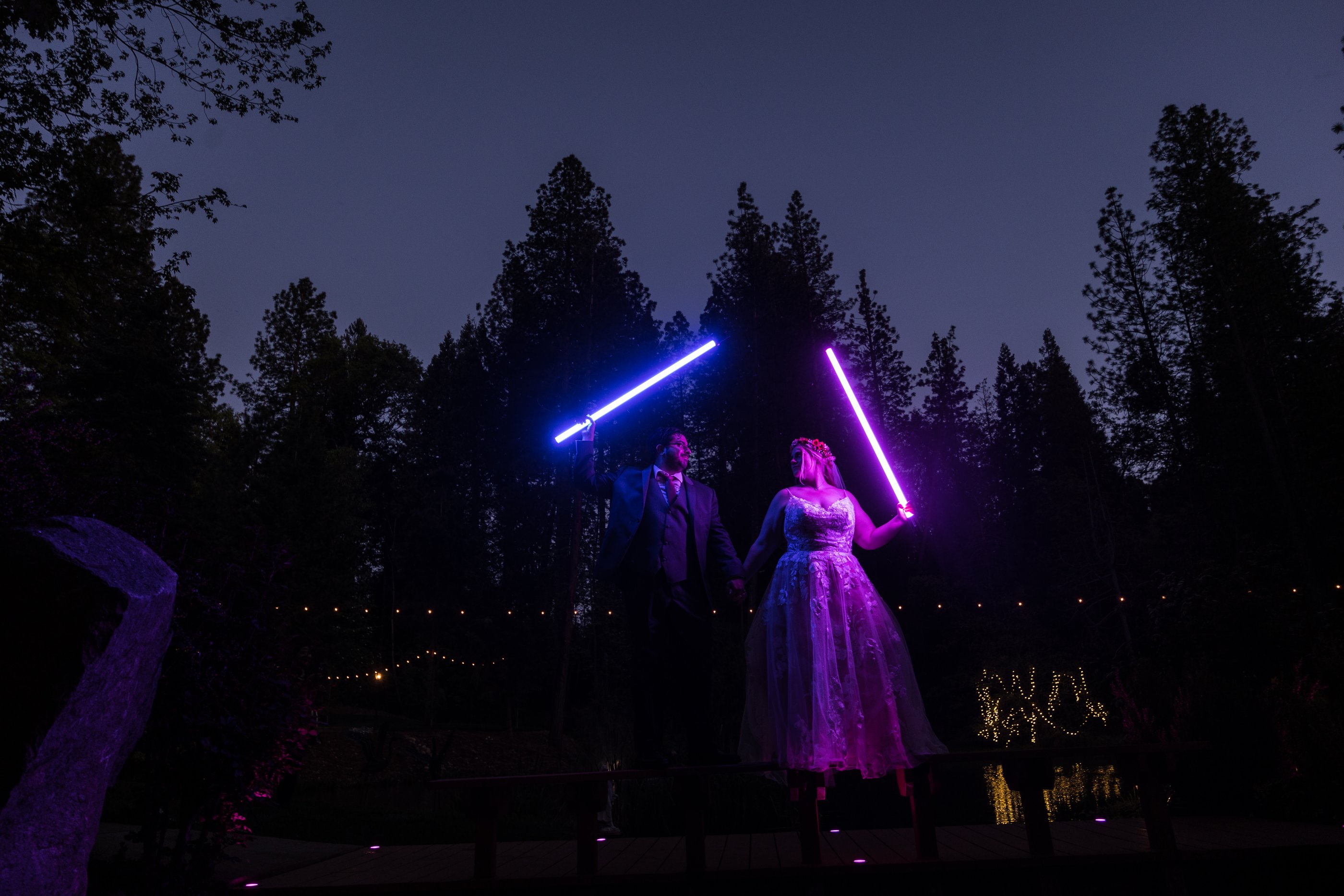 Abney and Chris raise glowing blue and purple lightsabers under the pines at dusk at Silverthorn Meadows