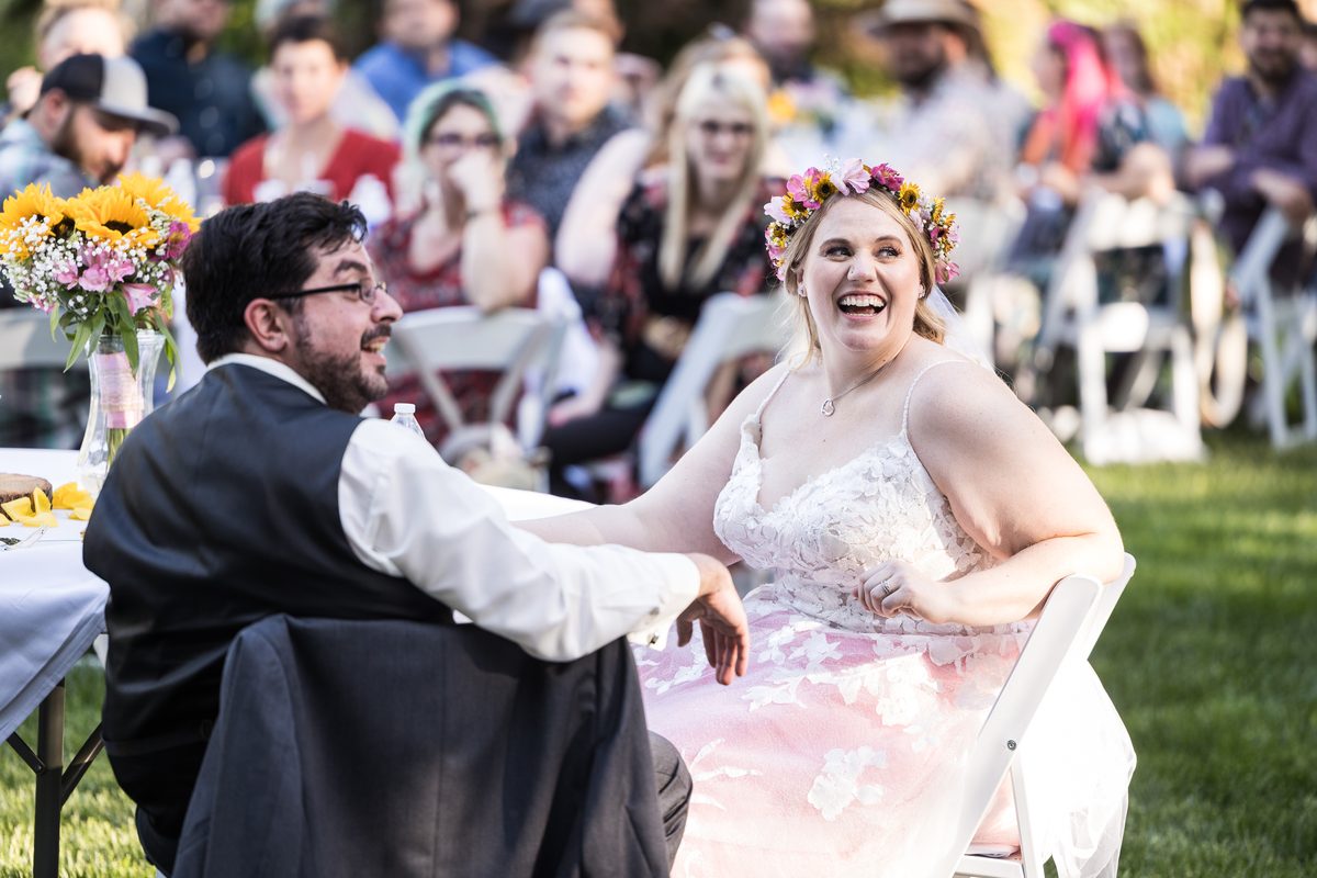 Abney laughs during the toasts, seated beside Chris at their table on the lawn with guests behind them