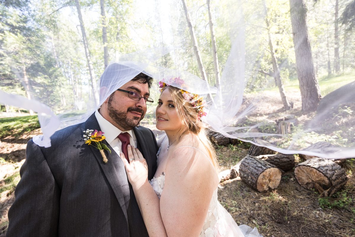 Abney and Chris stand close under her veil as it blows across the pine trees behind them