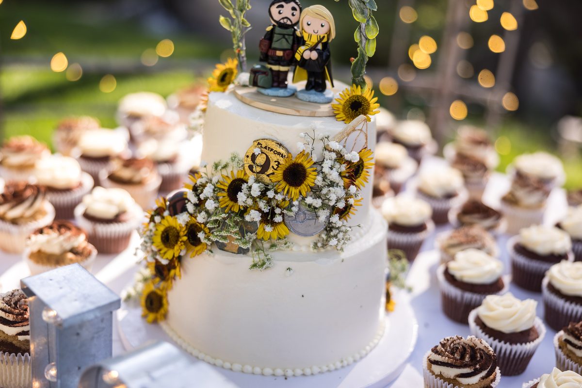 A white wedding cake trimmed with sunflowers and topped with two handmade figures of the couple, surrounded by cupcakes