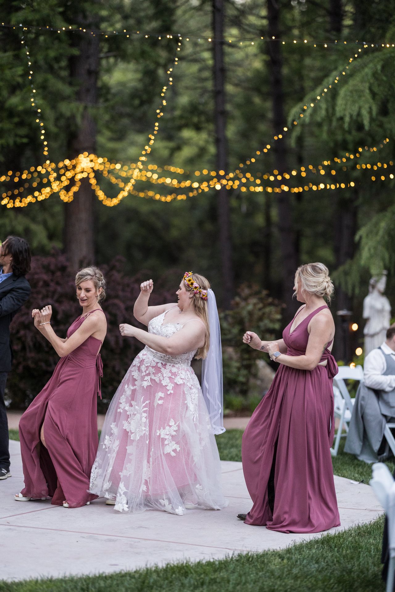 Abney dances between two bridesmaids in mauve dresses under a canopy of string lights