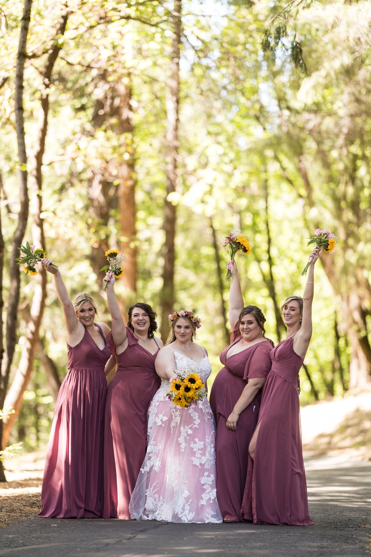 Abney and her bridesmaids raise their bouquets in the air on a forest path