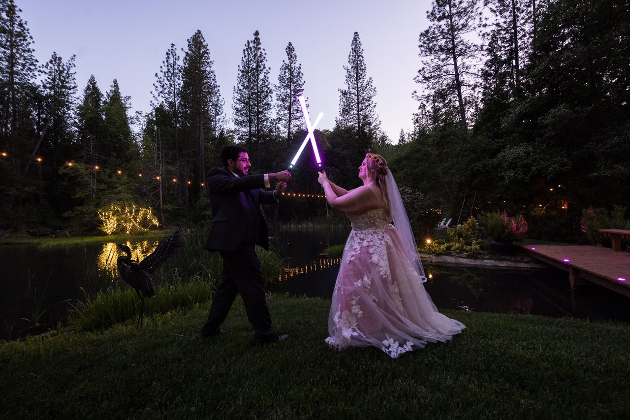 Abney and Chris cross lightsabers on the lawn beside the pond at dusk