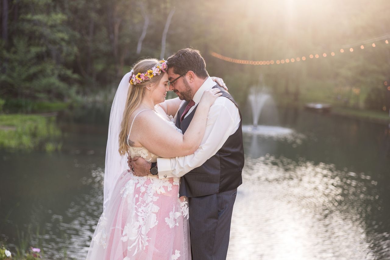 Abney and Chris rest their foreheads together beside the pond, with the fountain and string lights behind them