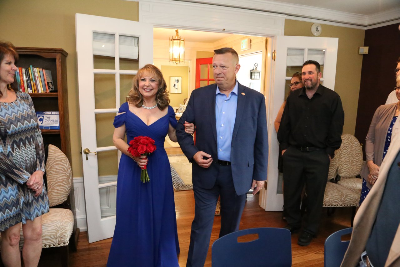 Alexanne walks in on the arm of a man in a blue suit as guests look on