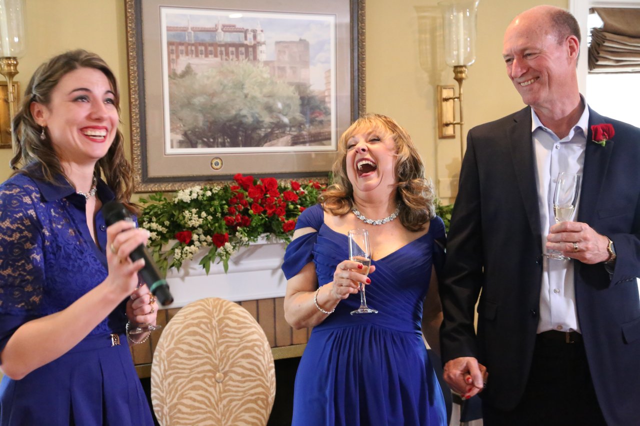 A young woman in blue lace gives a toast as Alexanne laughs and Tim smiles