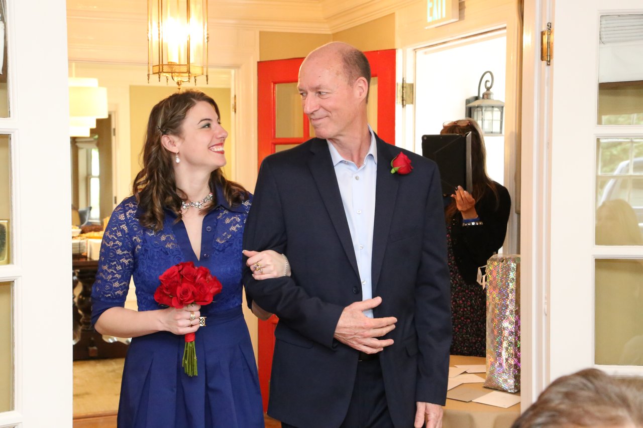 Tim walks in arm in arm with a young woman in blue lace carrying red roses