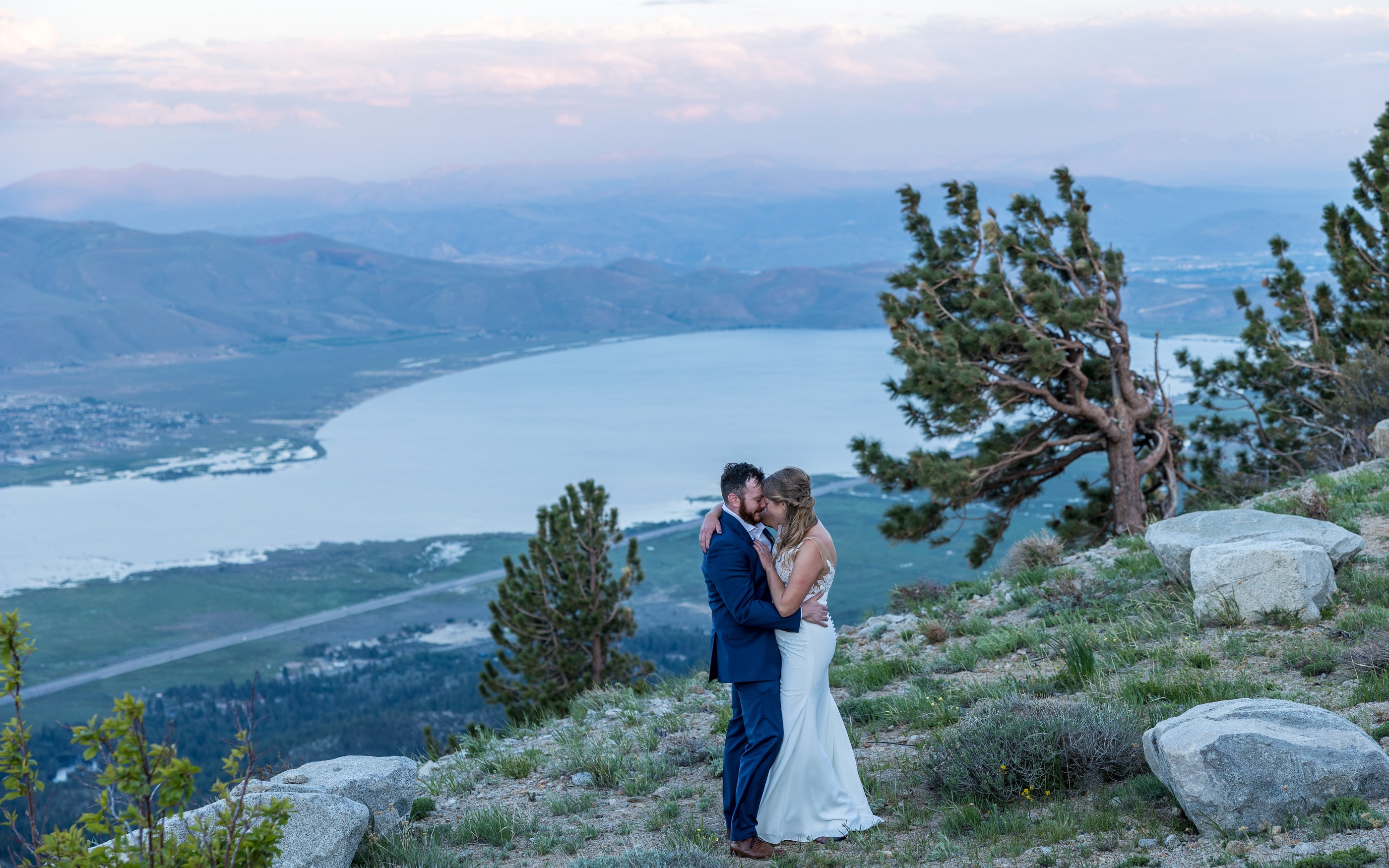 Ali and Robert kiss on the ridge at sunset beside a windswept pine, Washoe Lake far below