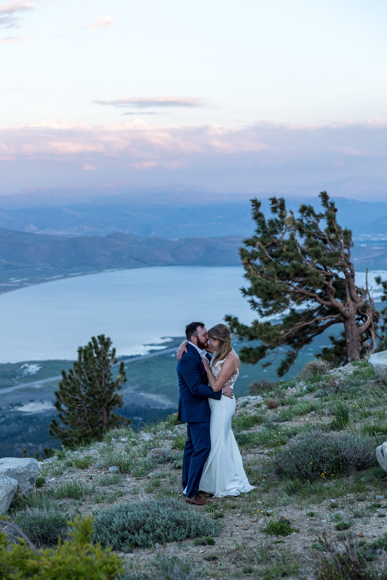 Ali and Robert kiss under pink clouds beside a windswept pine above Washoe Lake