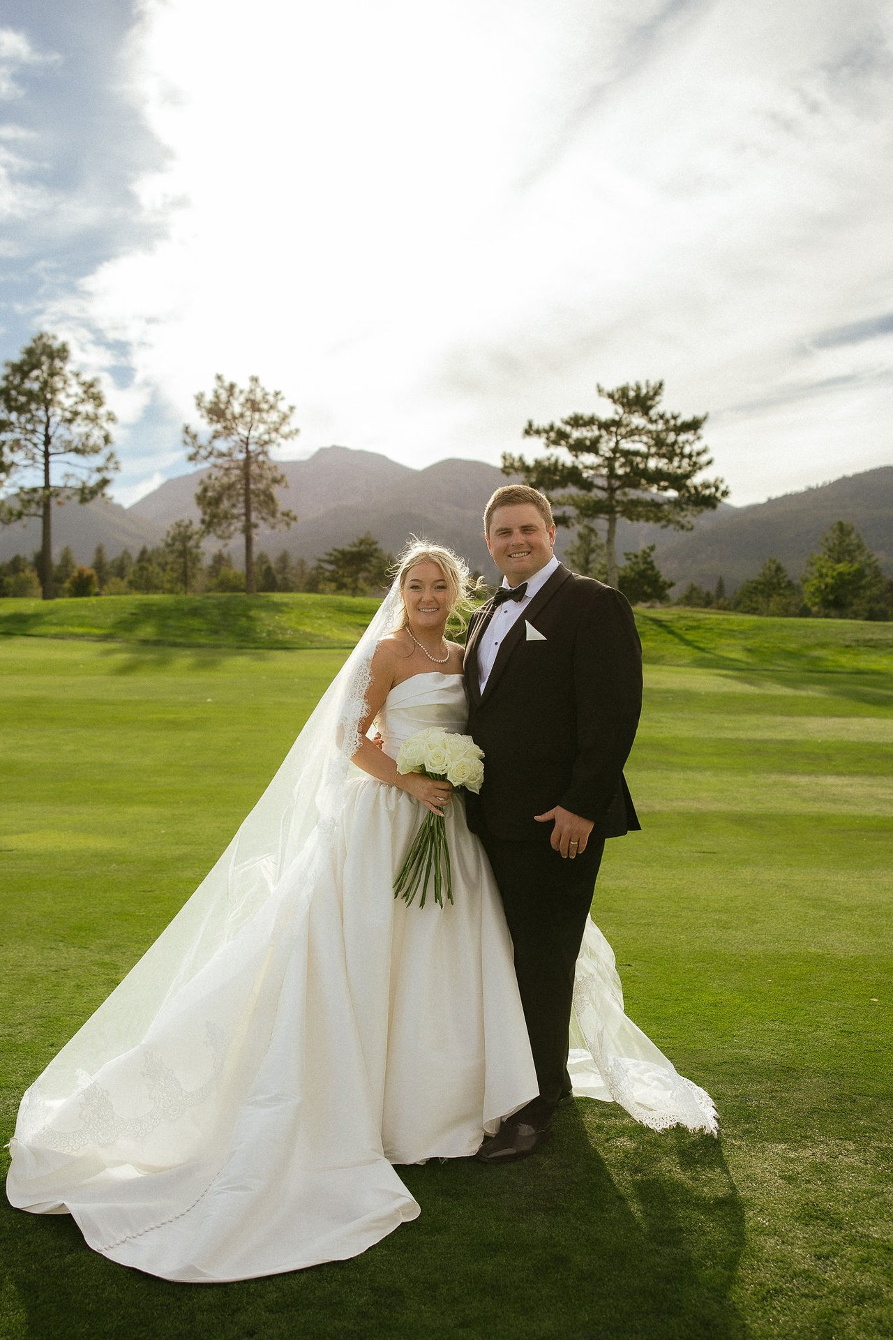 Ally and Drew stand together on the fairway, her veil and train across the grass, the pines and mountains behind them