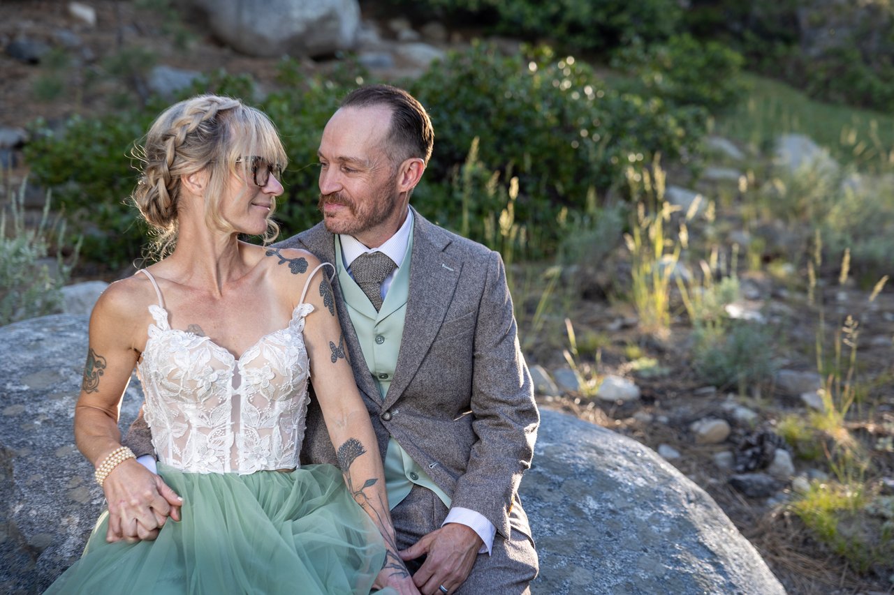 Amy and Justin smile at each other on a granite boulder in the forest