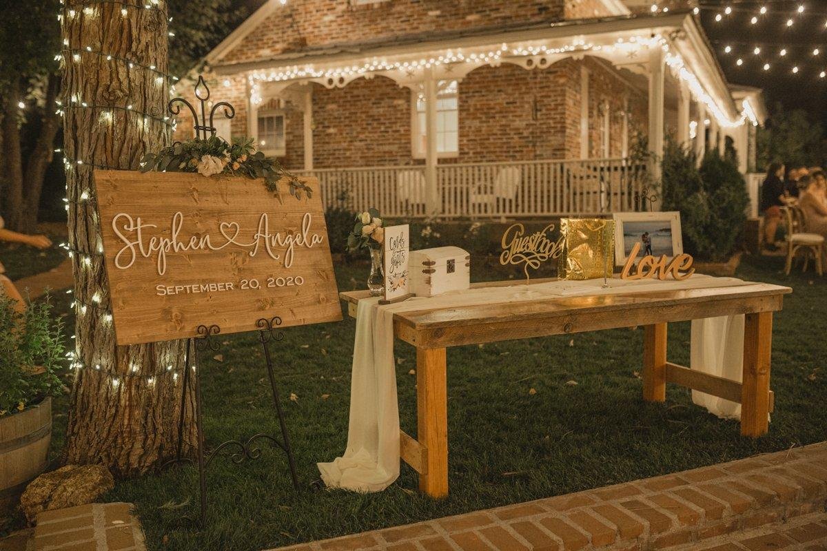 A wooden Stephen and Angela sign by the guest book table under the lights, photo by Composing Reality Photography