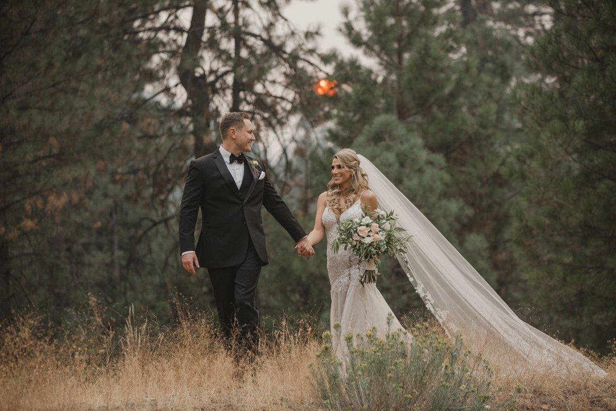 Angela and Stephen walk hand in hand through a meadow, her long veil trailing behind, photo by Composing Reality Photography