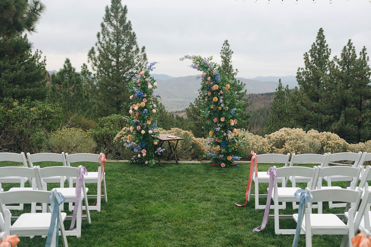 White chairs tied with ribbons face the floral arch on the lawn at the Tannenbaum Event Center, with pines and the valley beyond
