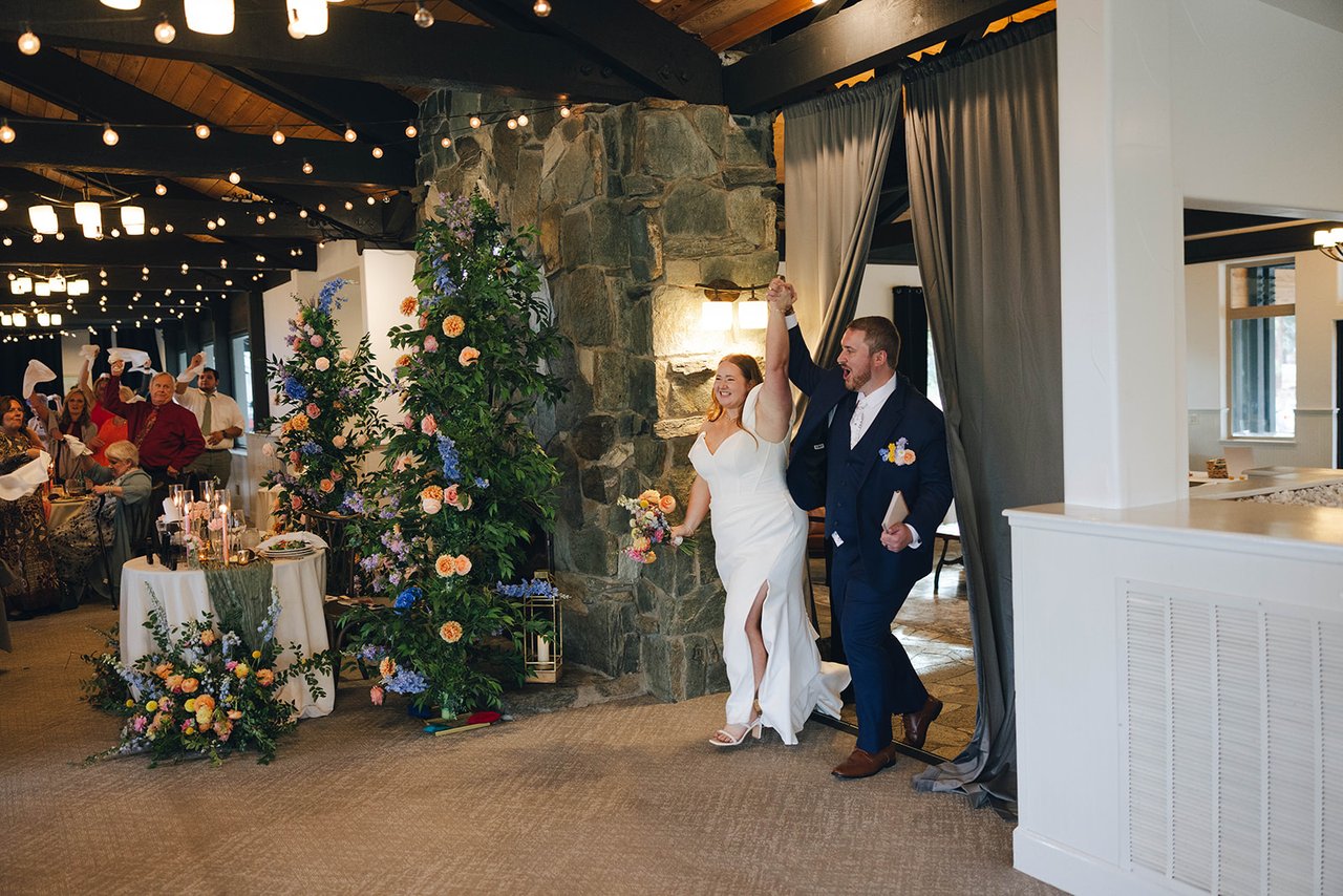 Autumn and Peter walk into the reception with arms raised, past the stone fireplace
